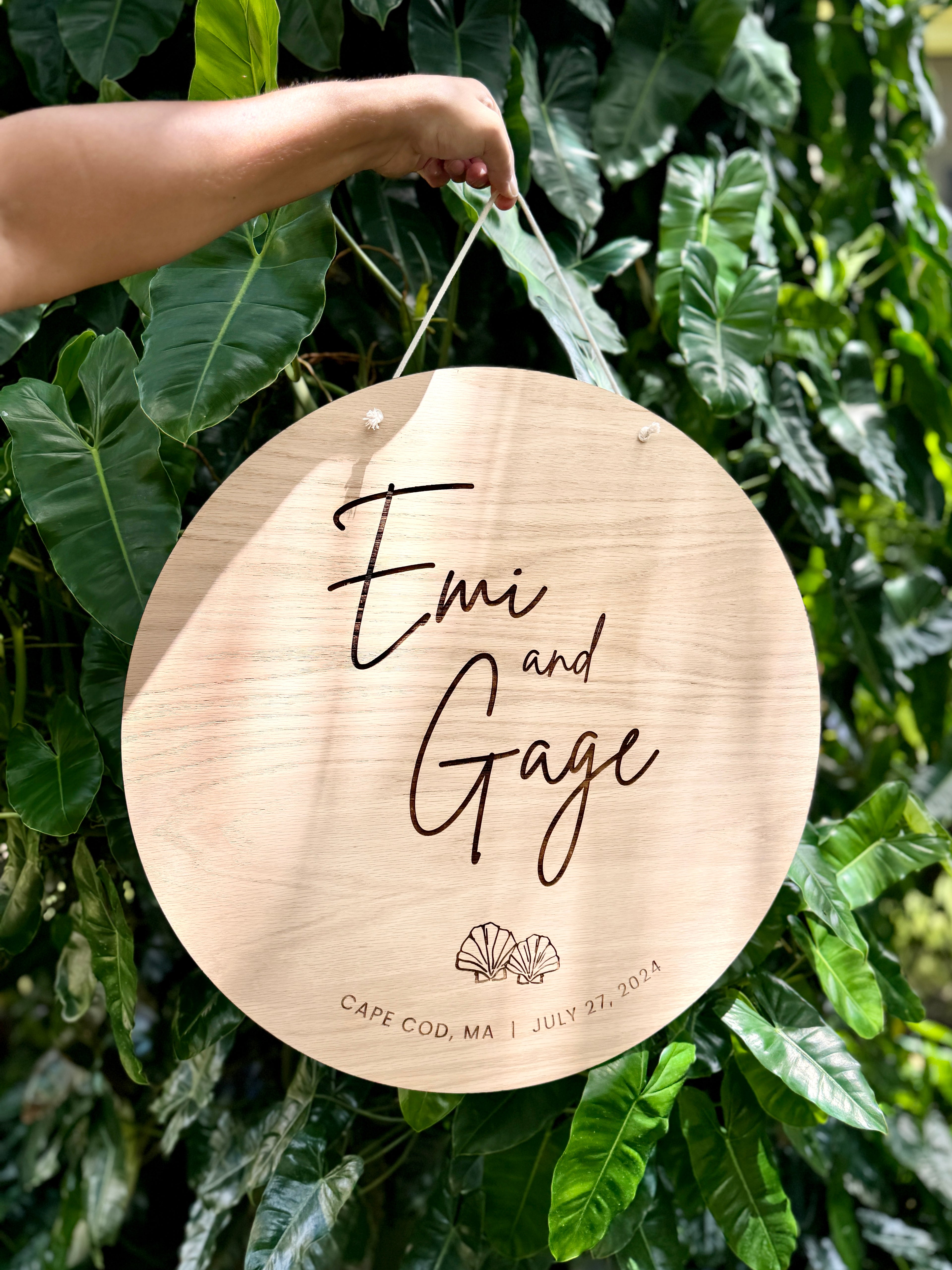 Wooden plaque with 'Emi and Gage' engraved, hanging against a green leafy background